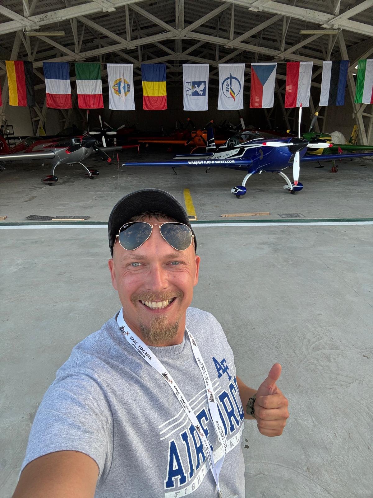 Maciej Kulaszewski w hangarze EAAC 2026 w Deva, za nim Extra 300L SP-EKS i flagi ekip