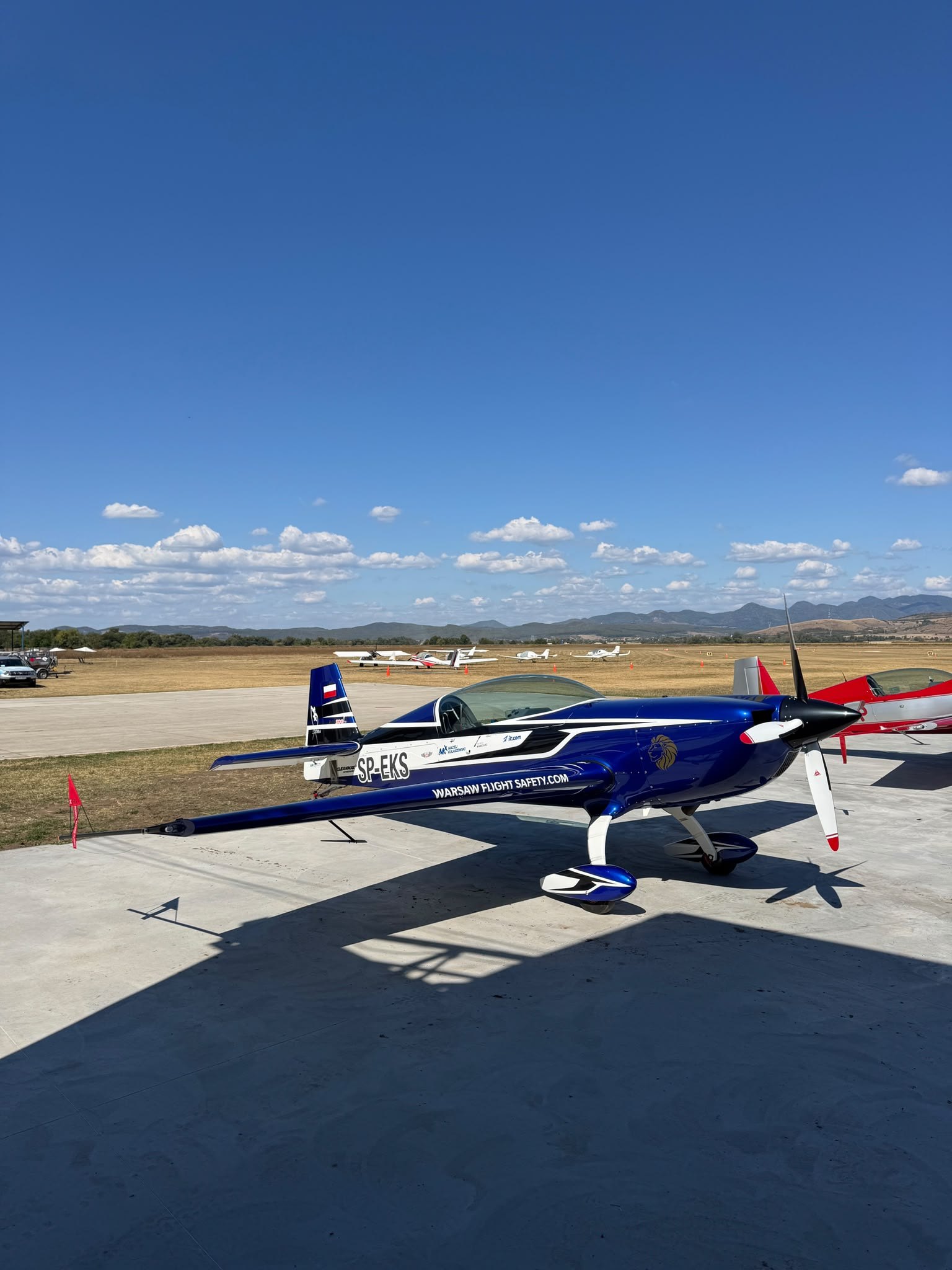 Extra 300L SP-EKS na płycie lotniska Deva/Săulești, w tle inne samoloty akrobacyjne i góry