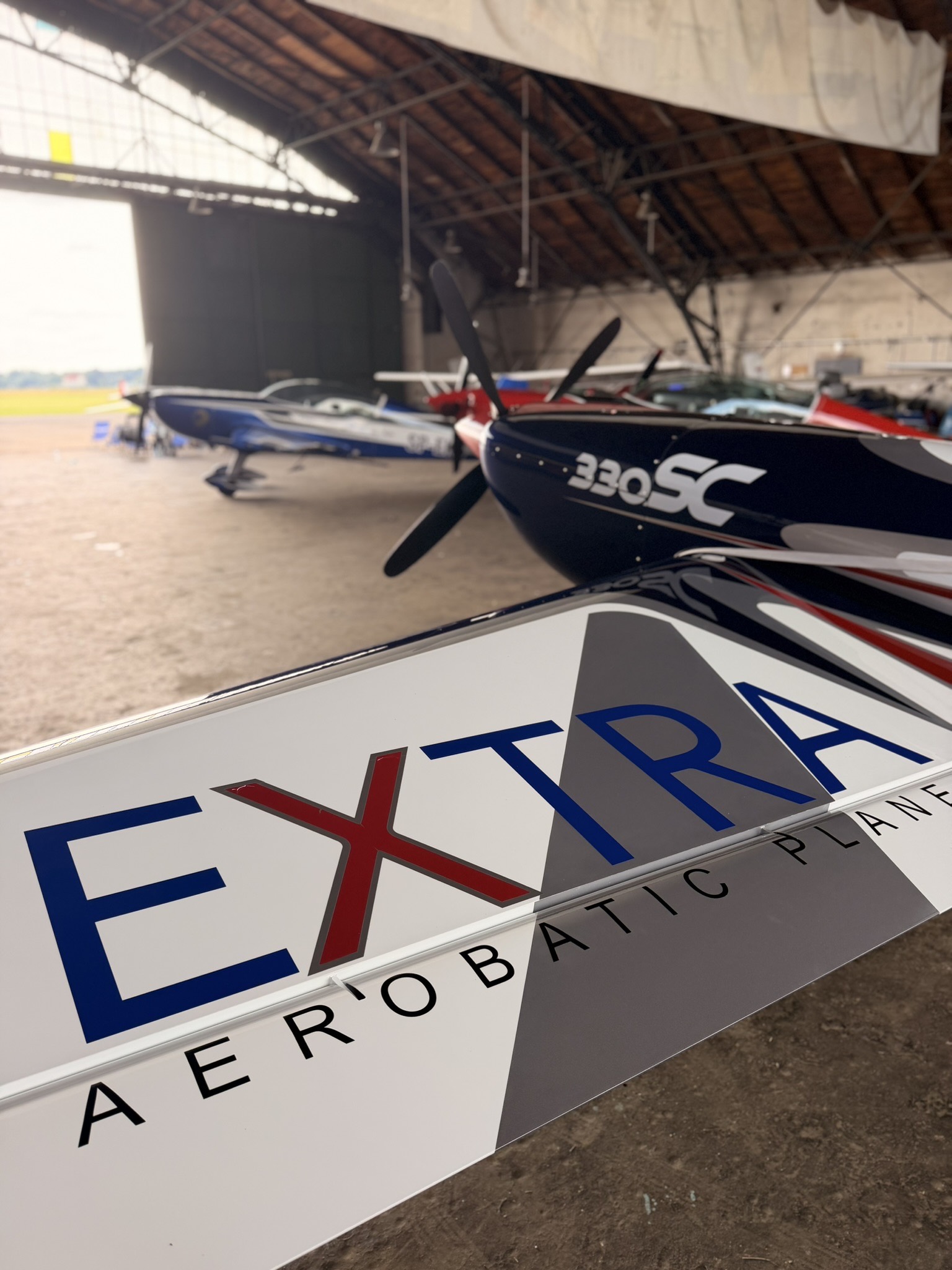 Skrzydło Extra 300L z logo Extra Aerobatic Planes, w tle samoloty akrobacyjne na 60. Mistrzostwach Polski w Pruszczu Gdańskim